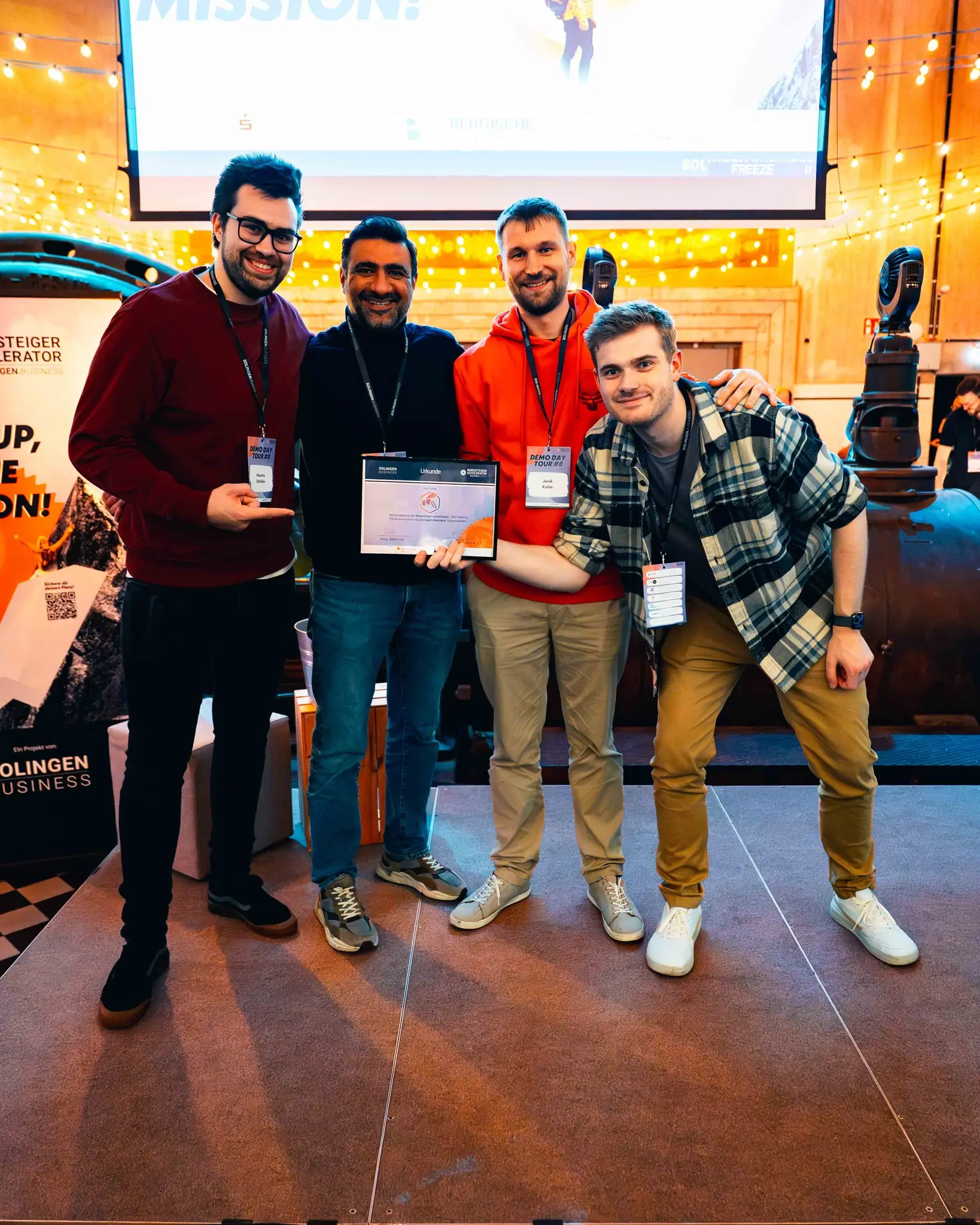 Vier Mitglieder des yourBeez-Teams stehen nebeneinander auf einer Bühne; in der Mitte halten zwei von ihnen eine gerahmte Urkunde des Bergsteiger-Accelerators, links steht ein Roll-up von Solingen Business, im Hintergrund hängen Lichterketten